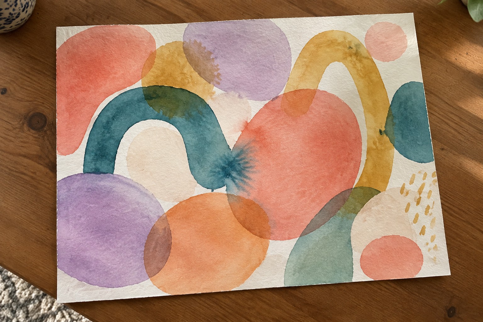 Abstract watercolor with colorful circles, arcs, and dots on white paper over wood.