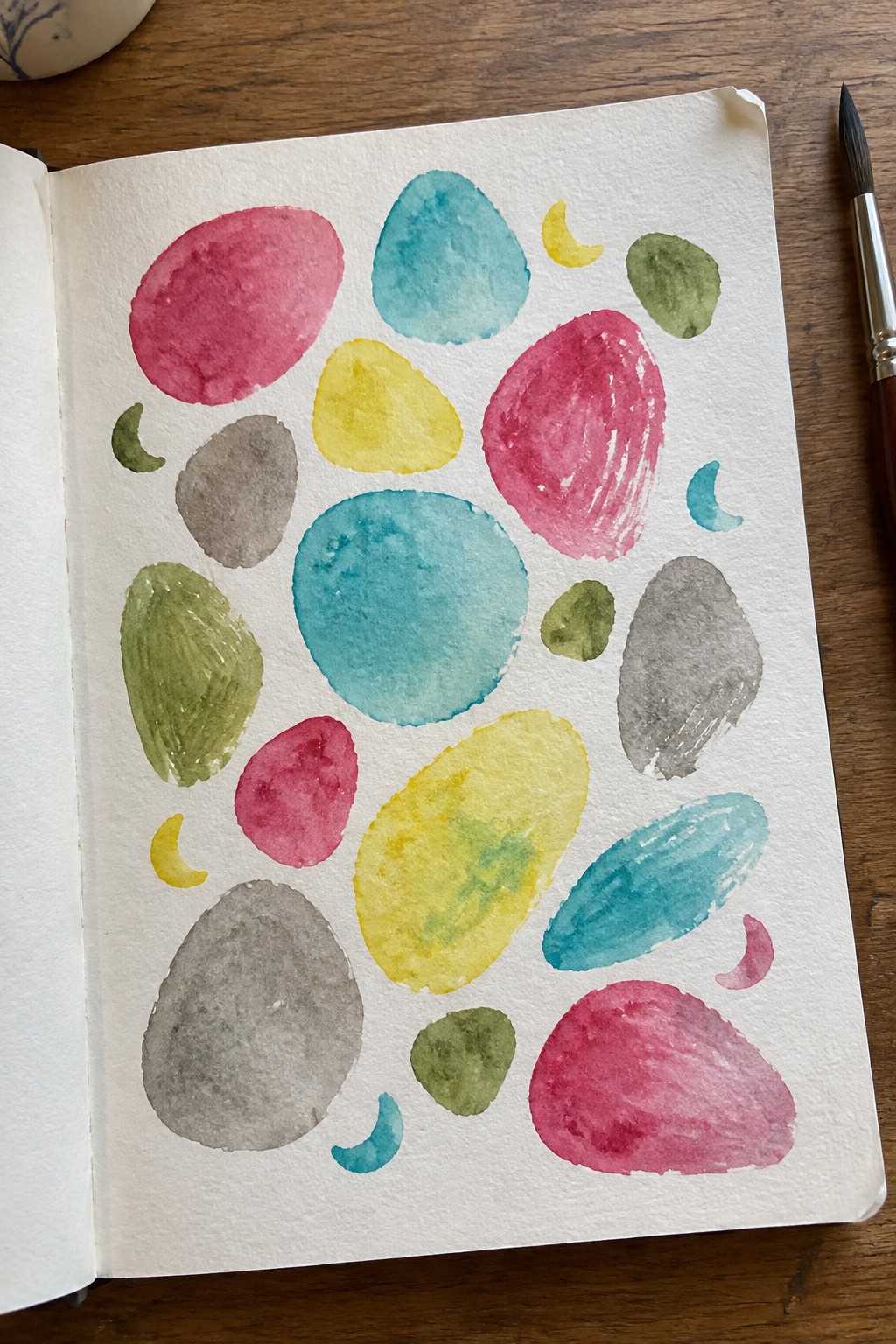 A sketchbook page with scattered watercolor shapes in pink, blue, yellow, green, and gray, including ovals and crescents.