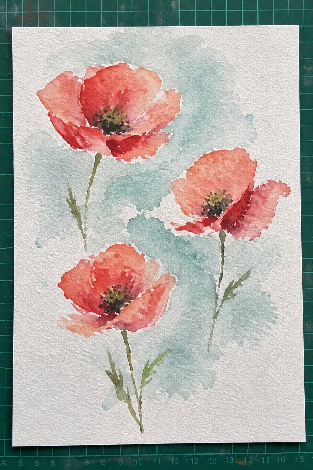 Watercolor painting of three red poppy flowers with green stems on white paper with light blue-green background washes.