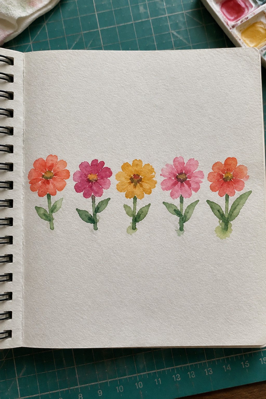Row of five colorful watercolor flowers with stems in open sketchbook on green mat.