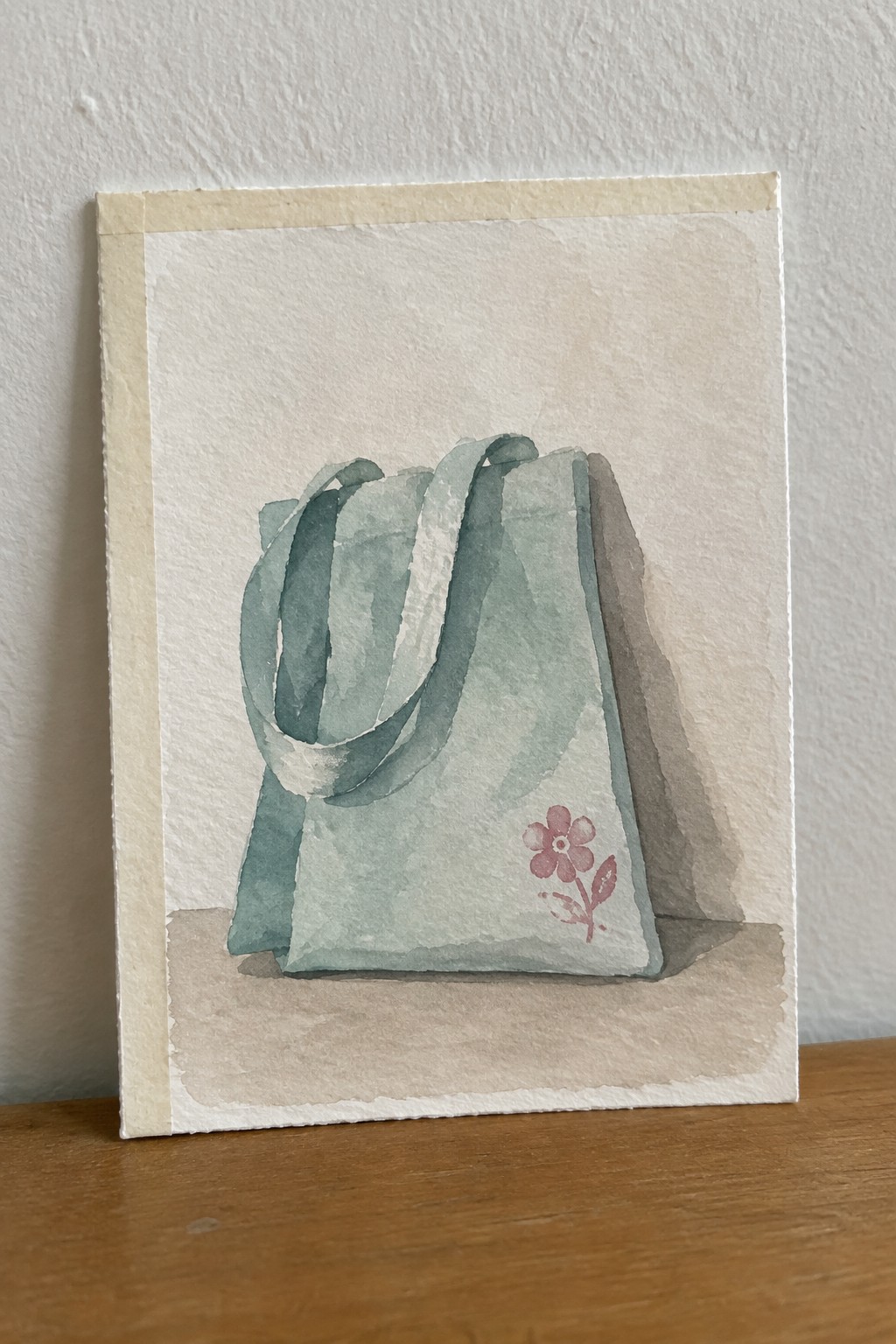 Watercolor of sage green tote bag with pink flower and soft shadow on wood