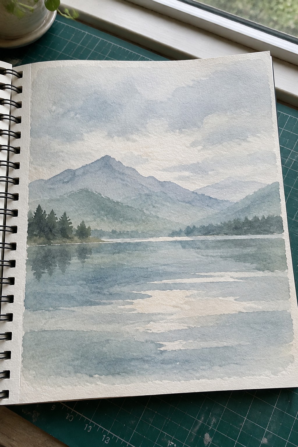 Watercolor painting of misty blue mountains reflected in a calm lake, open sketchbook on green mat