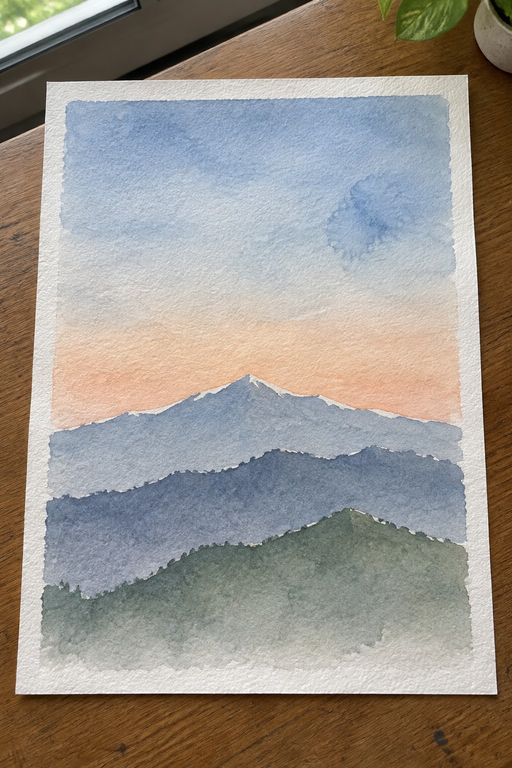 Watercolor landscape of layered blue mountains with green foreground under peach sky