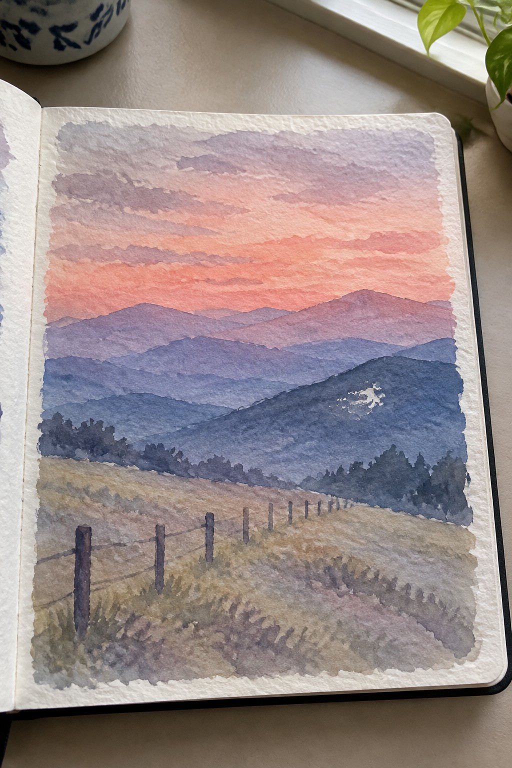 Watercolor painting in open sketchbook of sunset over layered mountains with fence