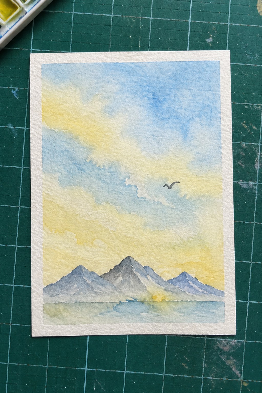 Watercolor landscape of mountains by water under blue-yellow sky with flying bird