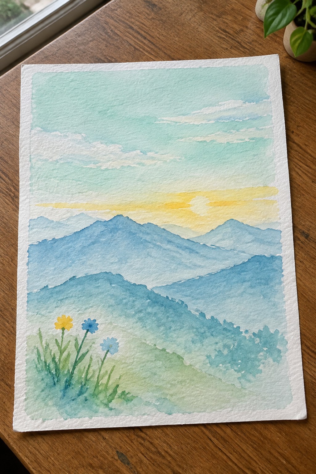 Watercolor of layered blue mountains under yellow sunset sky with blue and yellow wildflowers.