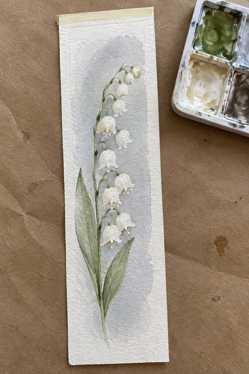 Watercolor bookmark of lily of the valley with white bell flowers and green leaves on a soft gray wash, shown next to a paint palette.