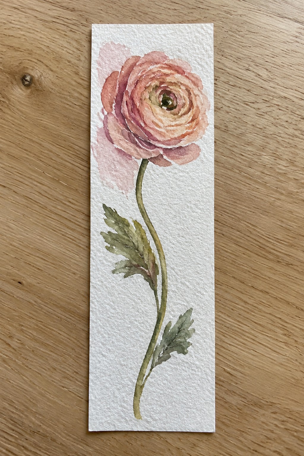 A watercolor painting of a pink and peach ranunculus flower on a long green stem with leaves, centered on a narrow white paper bookmark.