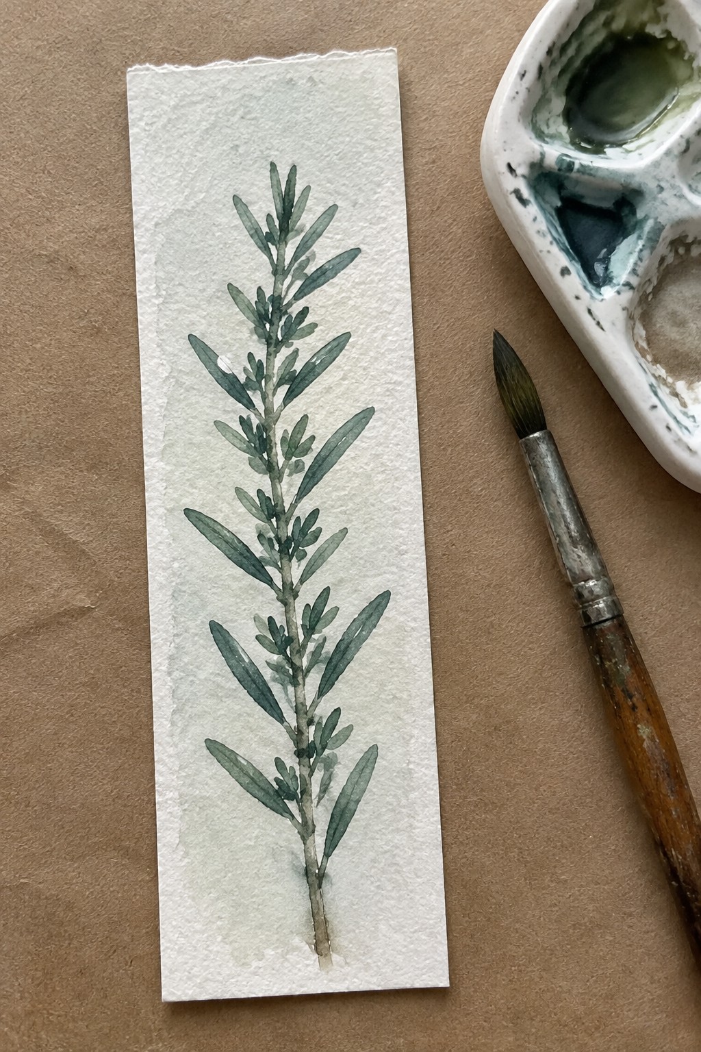Green watercolor branch on torn paper beside paintbrush and palette on brown surface.