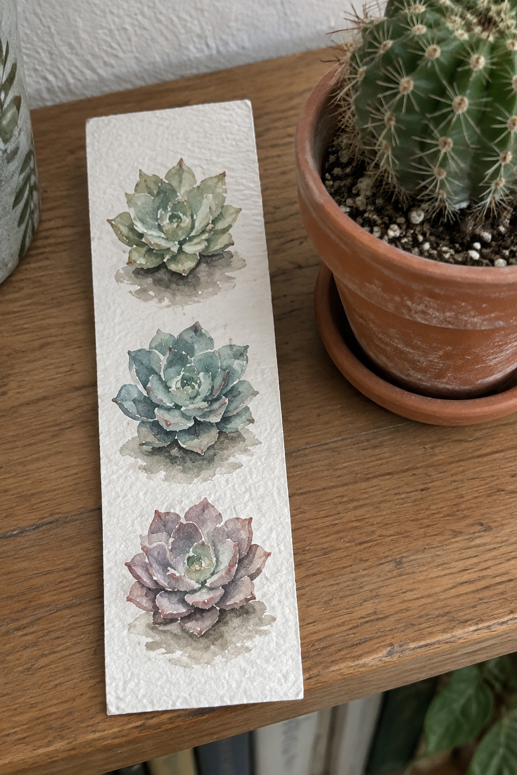 Three watercolor succulents on paper beside potted cactus on wooden table