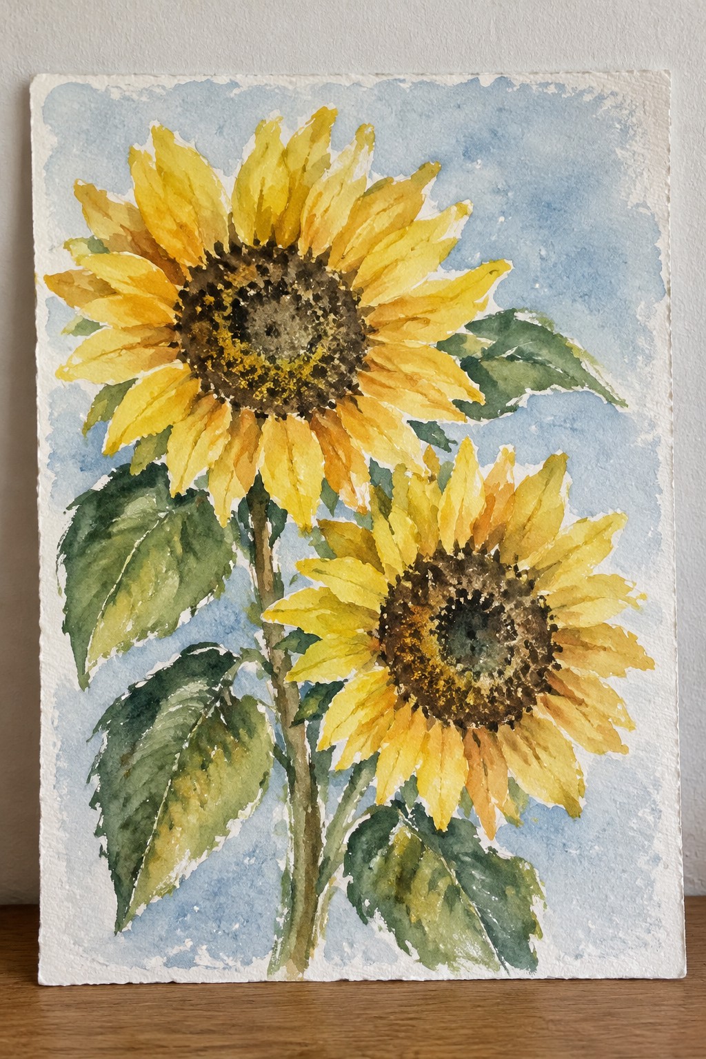 Two yellow sunflowers with green leaves in blue-sky watercolor painting.