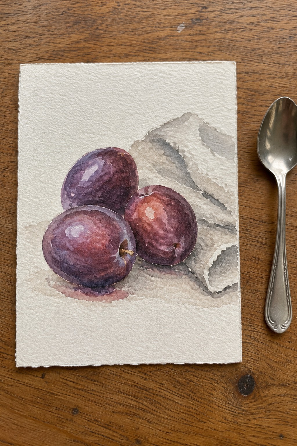 Three purple-red plums in watercolor on paper beside a metal spoon on wood