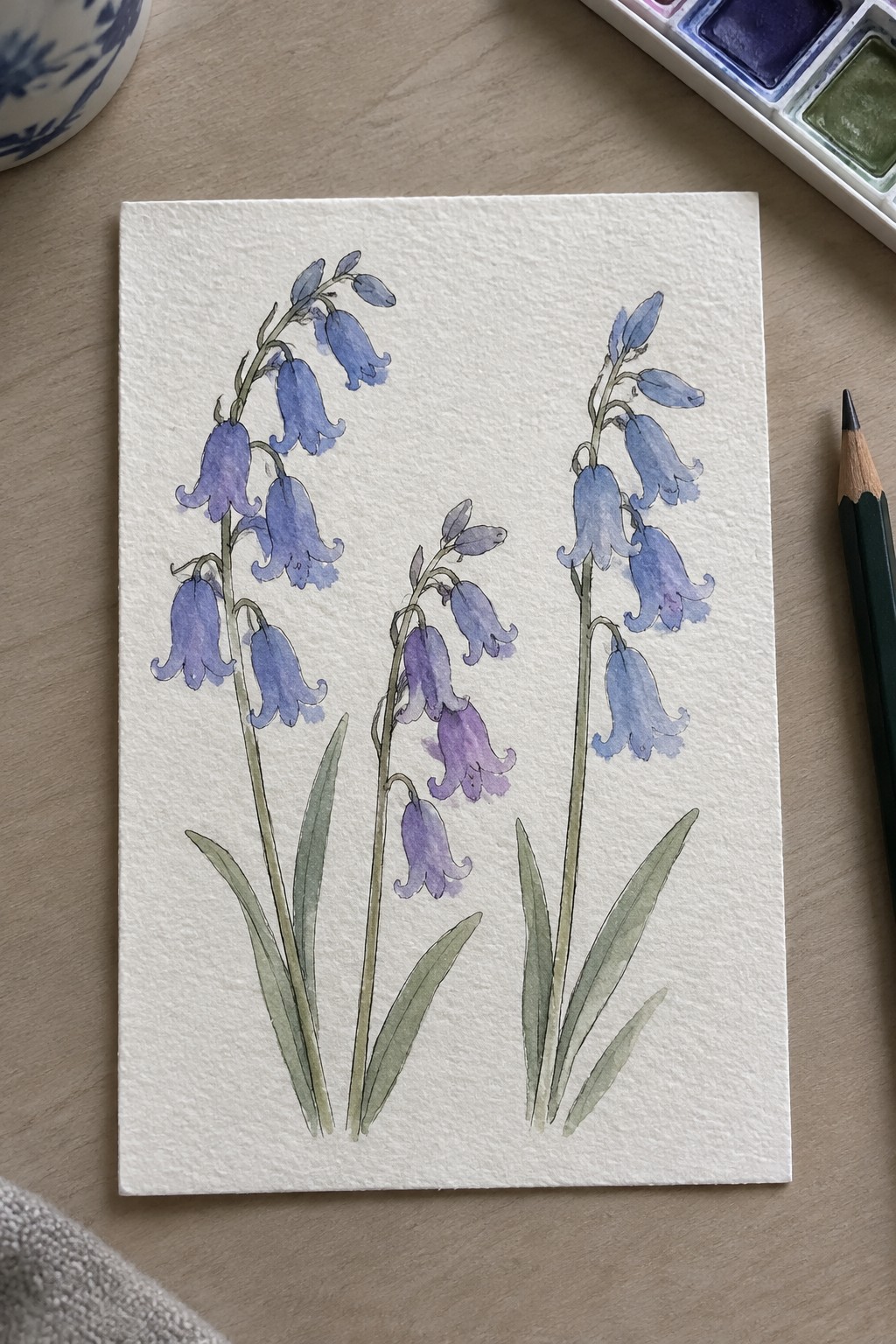 Watercolor bluebell flowers in purple hues with green stems on white paper beside palette and pencil