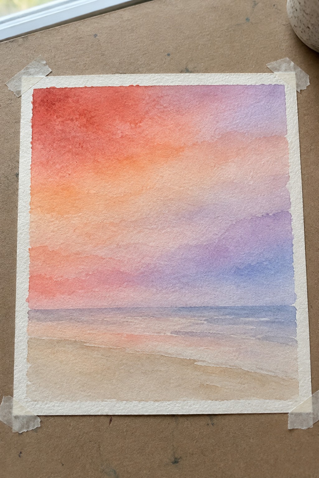 Watercolor sunset painting with red-orange-purple sky over beach horizon, taped to brown surface