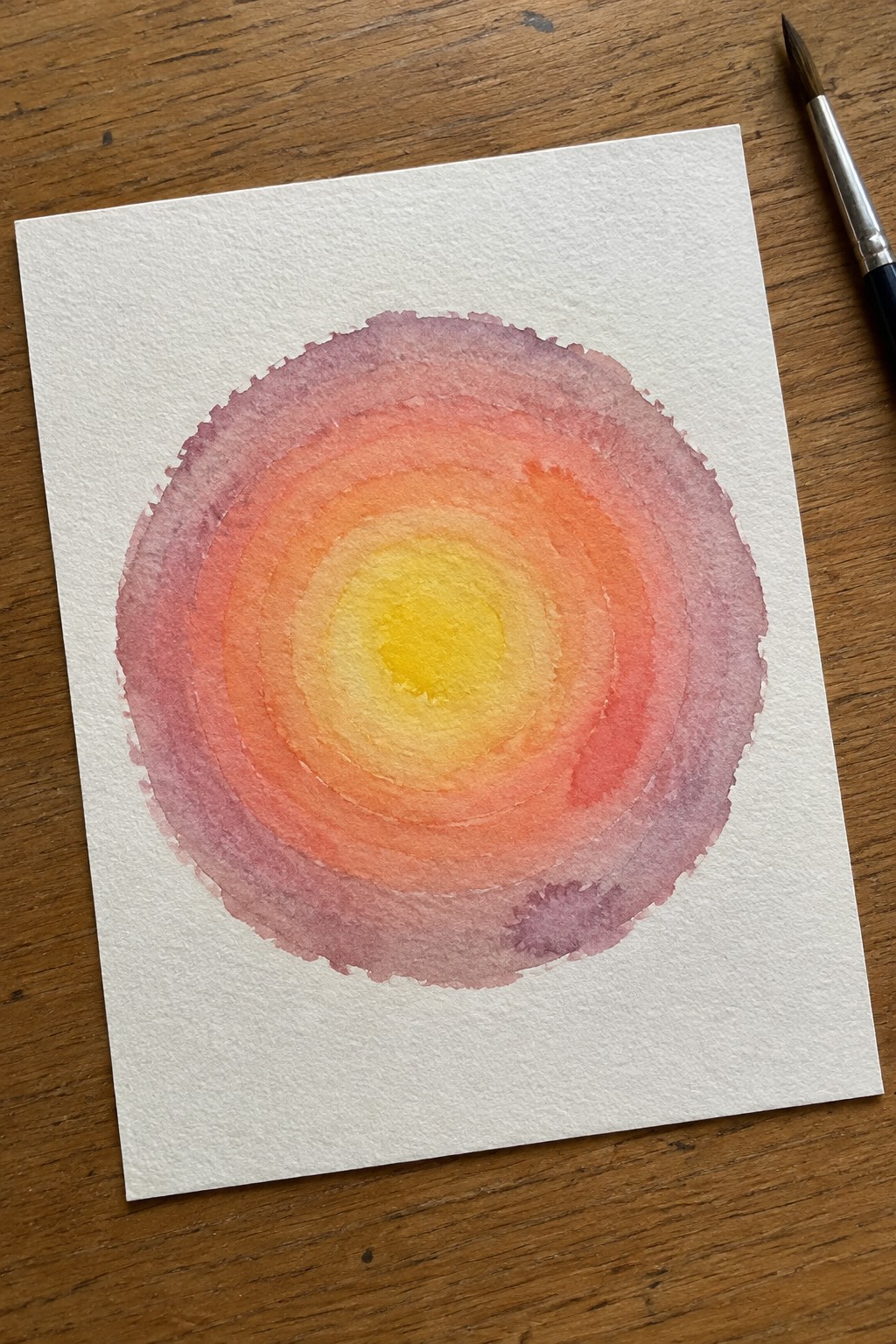 A loose watercolor painting of a circular gradient with yellow at the center blending outward through orange, pink, and purple rings on white paper, beside a paintbrush on wood.