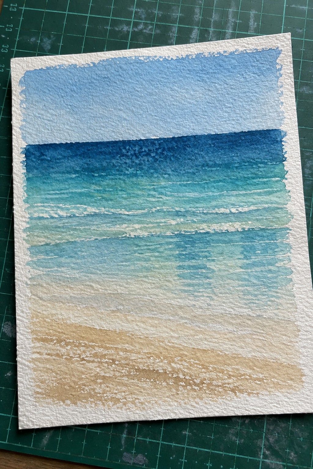 Watercolor seascape with blue sky, turquoise waves, and sandy beach on green mat