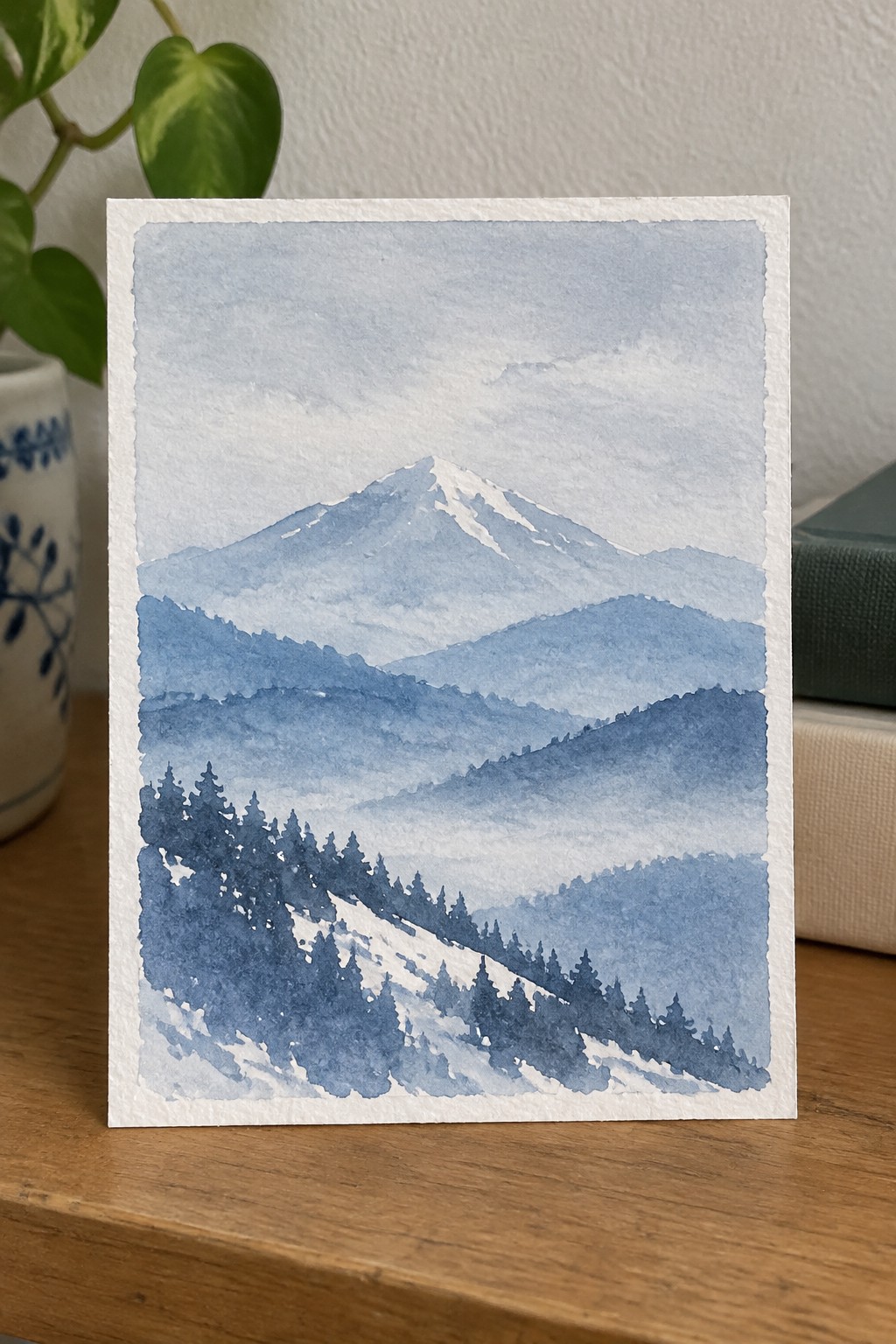 Blue watercolor landscape of snowy mountain peak above misty hills and pines