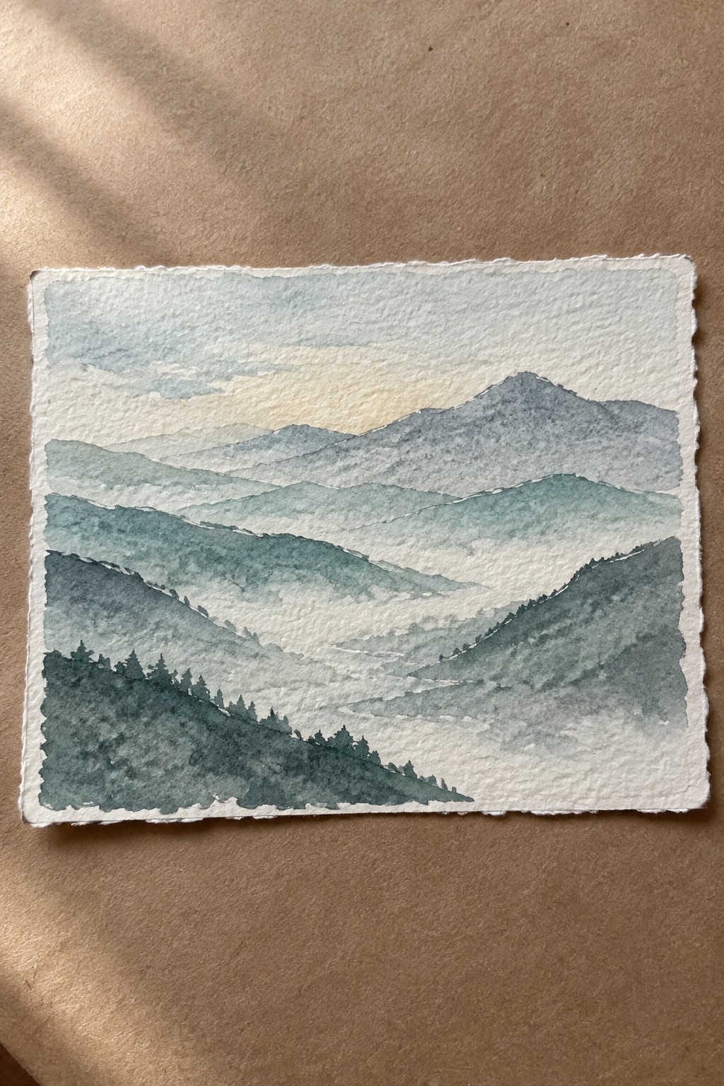 Watercolor of layered misty mountains in blue-green tones with pine trees at dawn