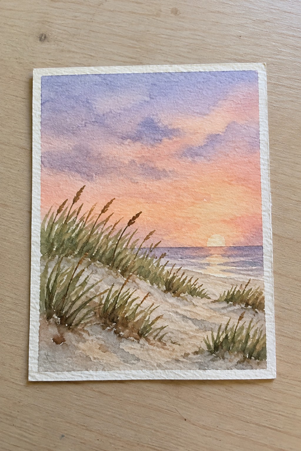 A watercolor painting of a beach at sunset showing sand dunes with green and brown grass, calm ocean water, and a sun low on the horizon under a sky of purple, pink, and orange.