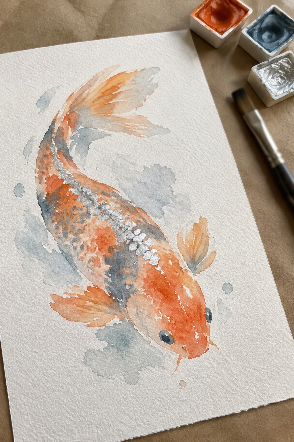 Watercolor koi fish in orange and gray with paint pans and brush nearby