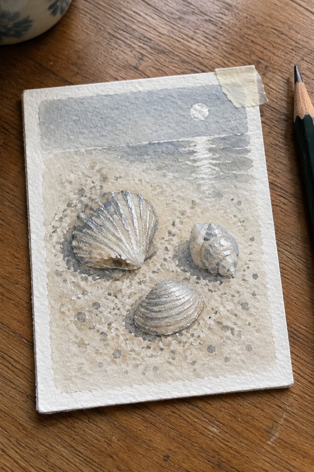 Three metallic seashells on watercolor beach scene with gray horizon and pencil nearby