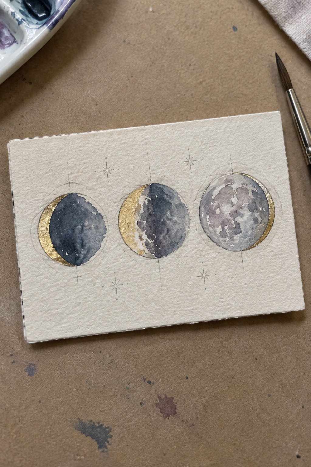 Watercolor painting of three moon phases with gold edges and sketched stars on paper