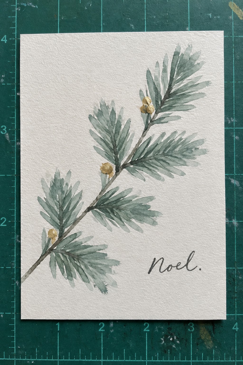 Watercolor evergreen branch with green needles, gold berries, and Noel script on white paper