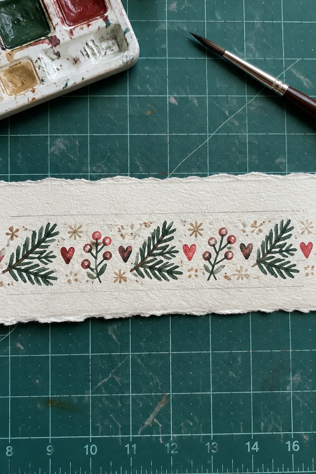 Watercolor palette, brush, and paper strip with green leaves and red hearts on mat