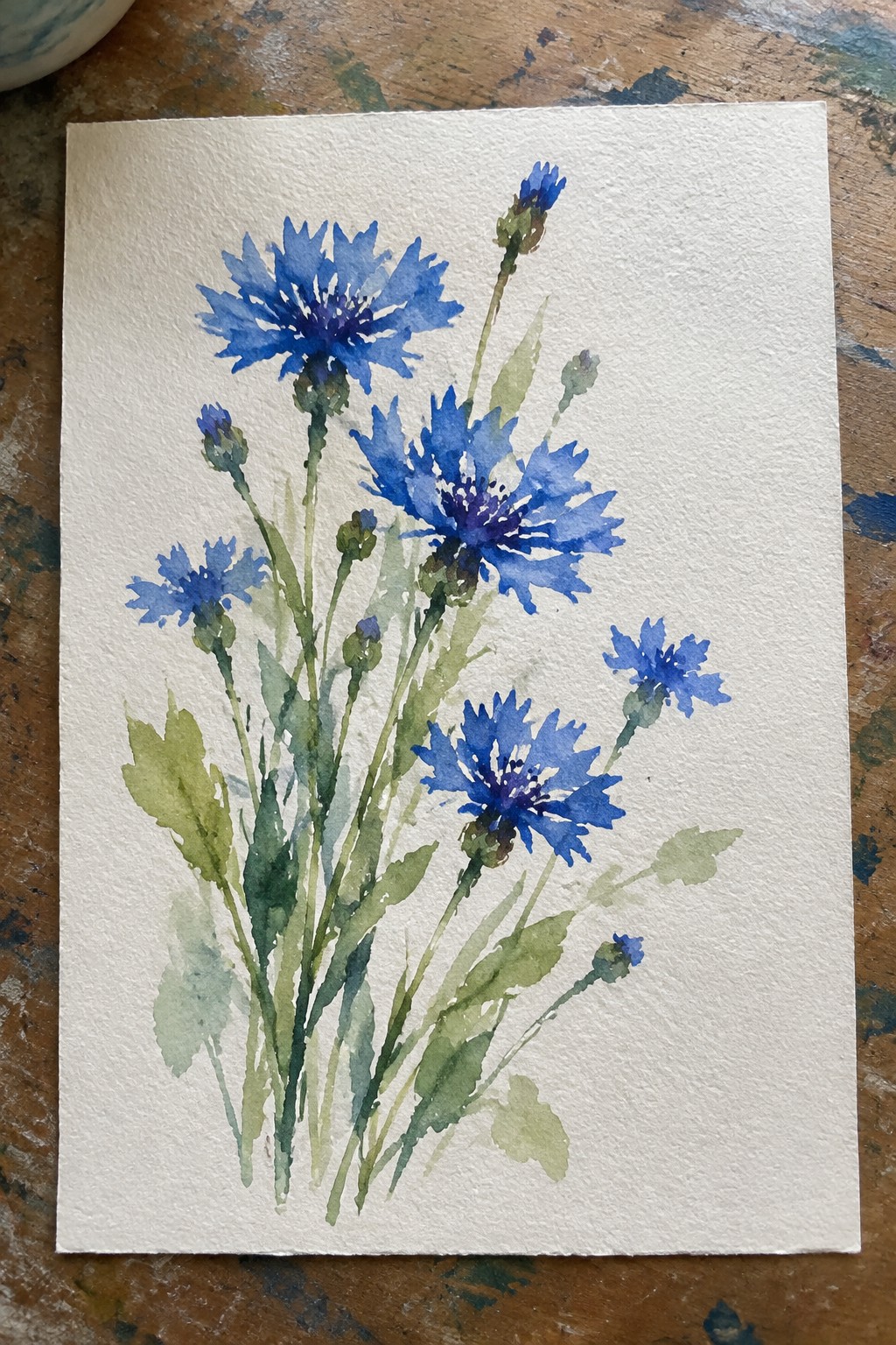 A loose watercolor painting of blue cornflowers with green stems and leaves on white paper.