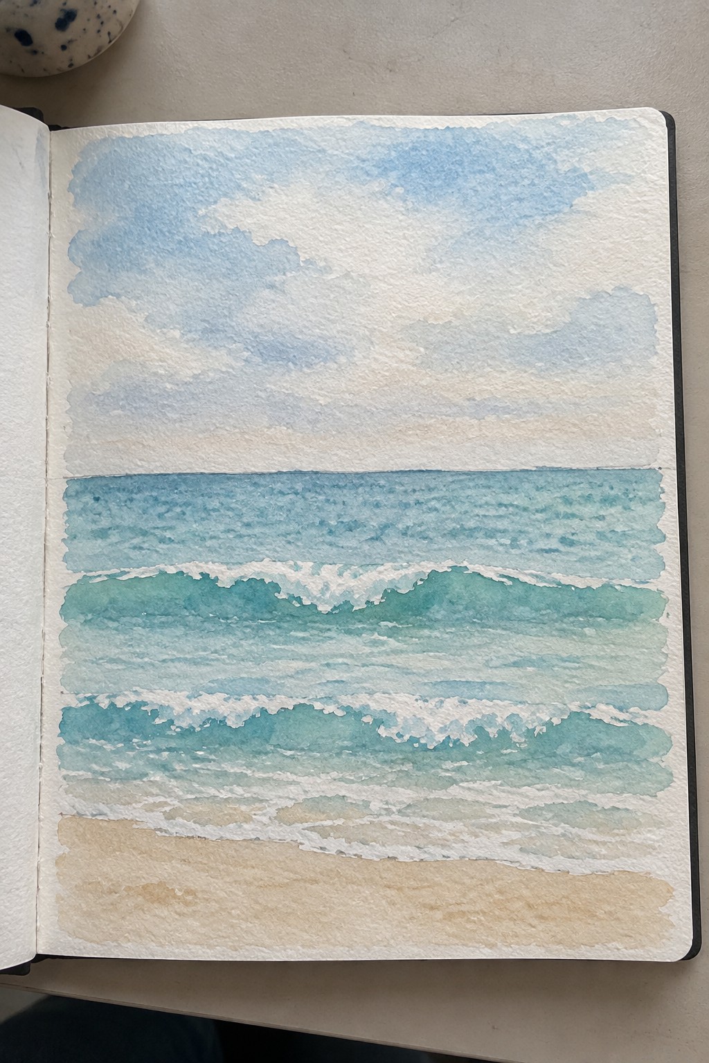 Open sketchbook showing watercolor of turquoise waves with white foam on sandy beach
